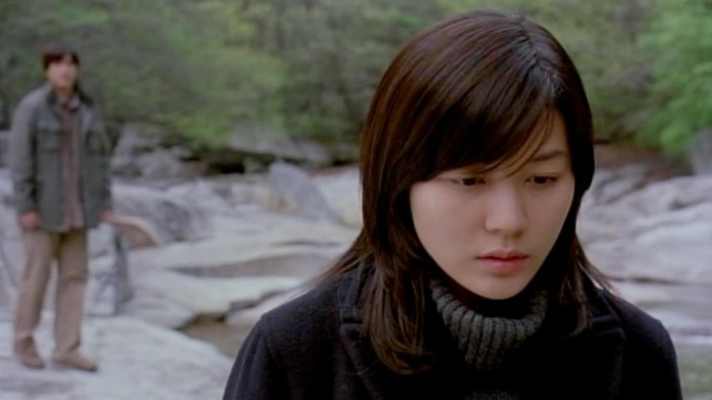 Min Ji-won (Kim Ha-neul) who suffers stands in a rocky forest while a man stands behind her in Dead Friend (2004)