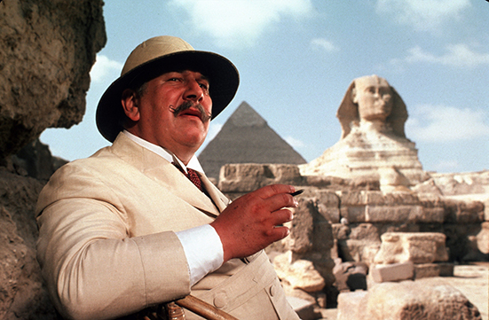 A man with a mustache in a white suit and hat stands with Egyptian monuments looming behind