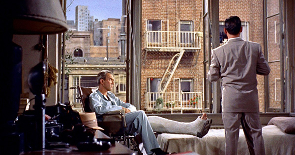 A man sits in his wheelchair as another man looks through an apartment's giant windows