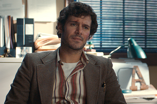A young man with black hair in a brown coat and striped shirt sits in an office