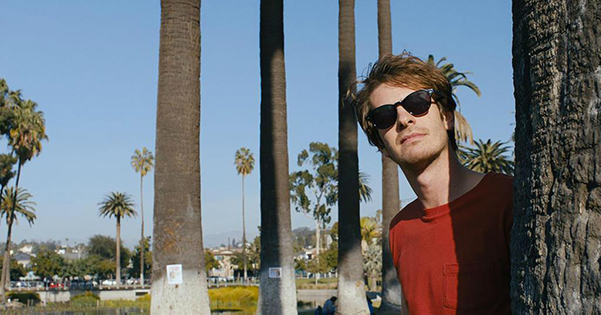 A young man in a red shirt wearing sunglasses looks from behind a palm tree