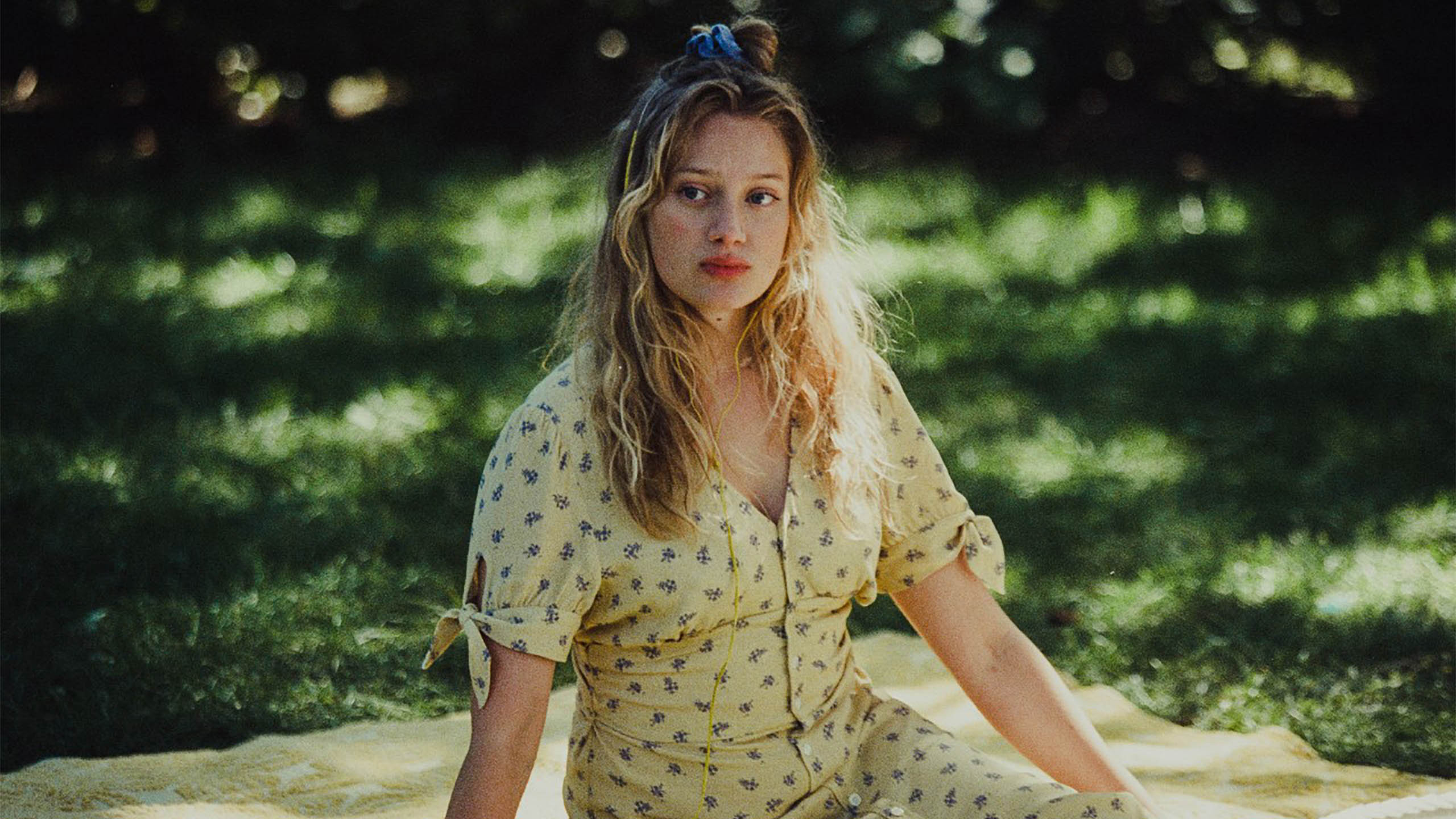 A blonde woman in a yellow dress sits in a blanket in the grass
