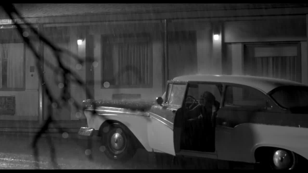 A black and white image of a car with its door open outside a motel
