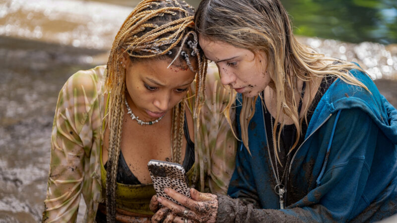 Twi girls drenched in water and sprinkled with dirt look down at a phone