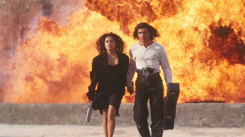 A man carrying a guitar case and a woman carrying a gun walk away from a wall of fire