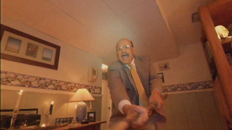 An older man in glasses wearing a suit swings a stick towards the camera
