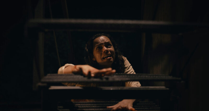 A women looking scared crawls up some stairs from a dark room