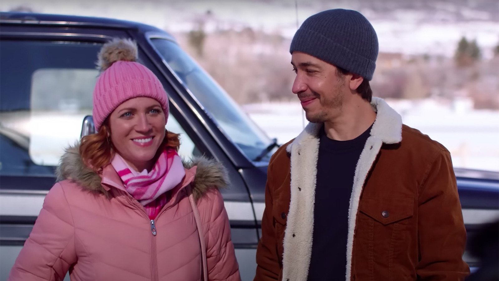 A woman in a pink outfit and a man in a read coat stand next to a truck in the snow