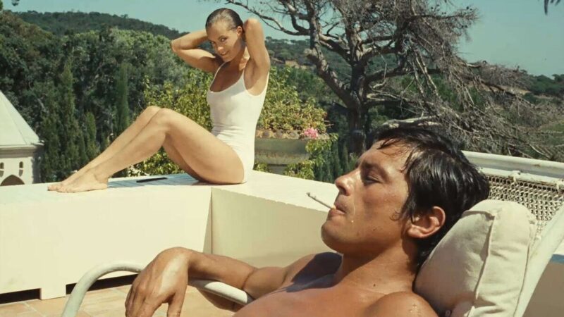 A man sits and smokes a cigarette as a woman in a white bathing suit stares at him