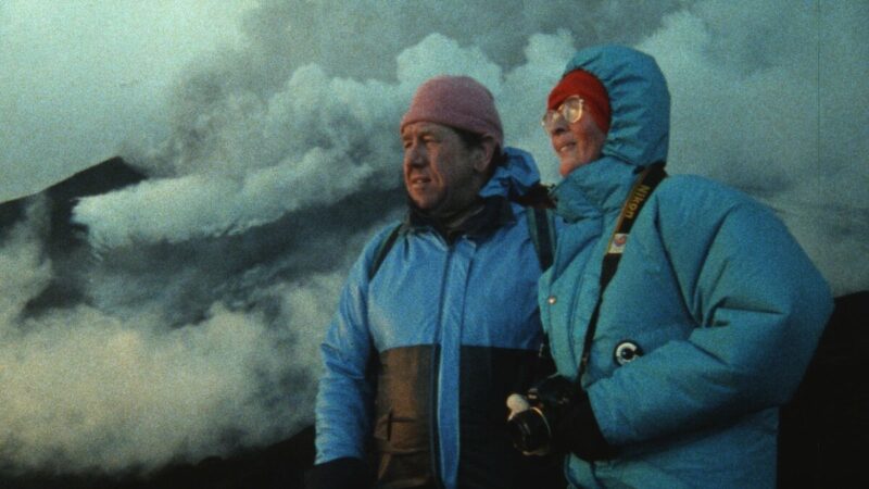A man and a woman wearing blue coats stand near a volcano as smoke floats by