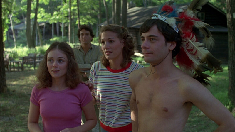 Four young adults stand facing the same director at a summer camp