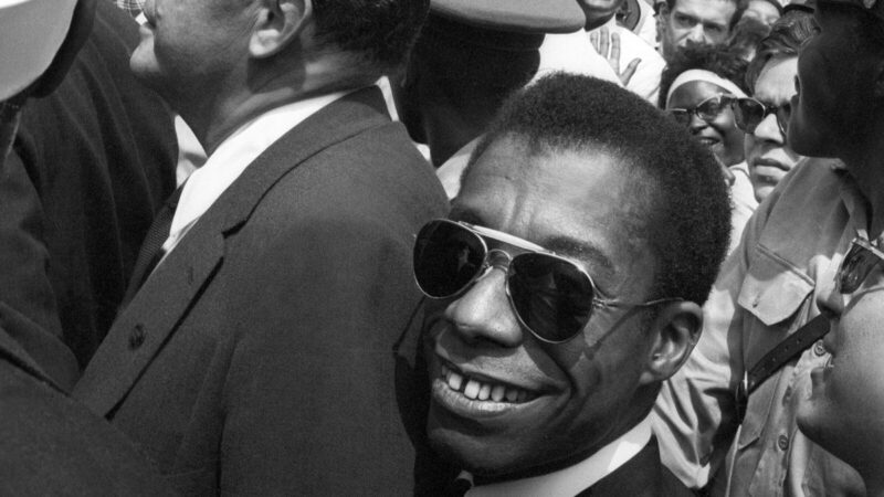 A man wearing sunglasses smiles while standing in a crowd