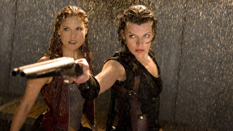 A woman stands to the left of another woman holding a shotgun as water falls from sprinklers
