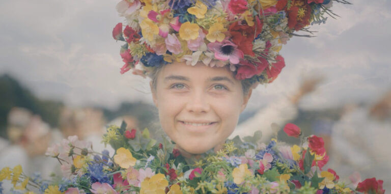 Dani wears a dress and crown of flowers and smiles as she gazes upon something.