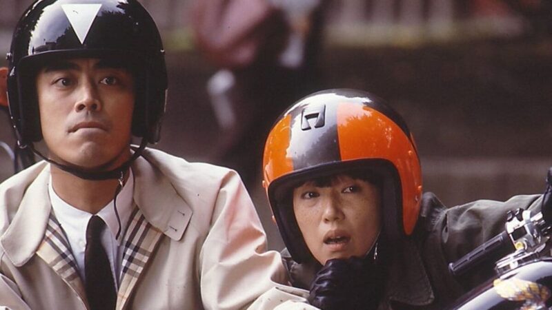 A man and a woman wearing motorcycle helmets stare at something in the distance