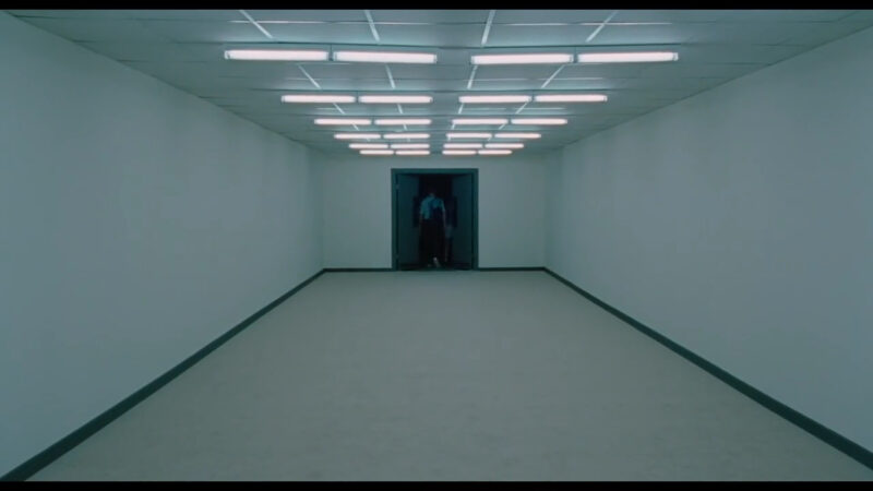 A man walks through a door at the end of a long hallway