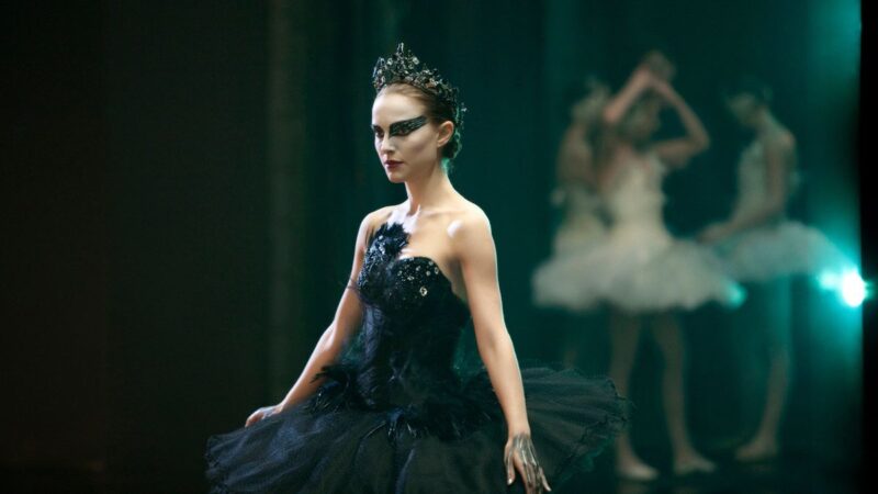 A ballerina dressed in black stands with other ballerinas dressed in white in the background