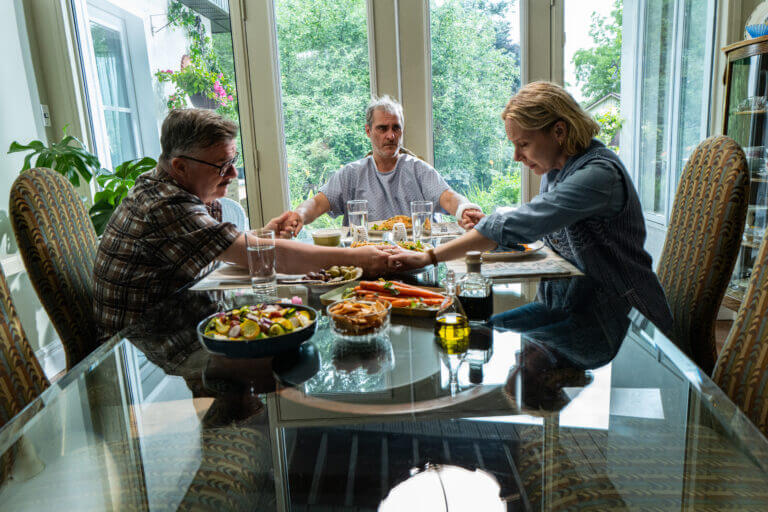Beau sits at a table with Roger and Grace. The three hold hands before a meal.