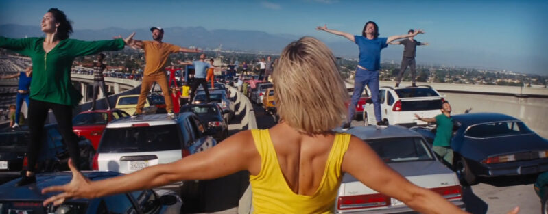 A woman in a yellow dress faces a number of people standing on their cars on a highway