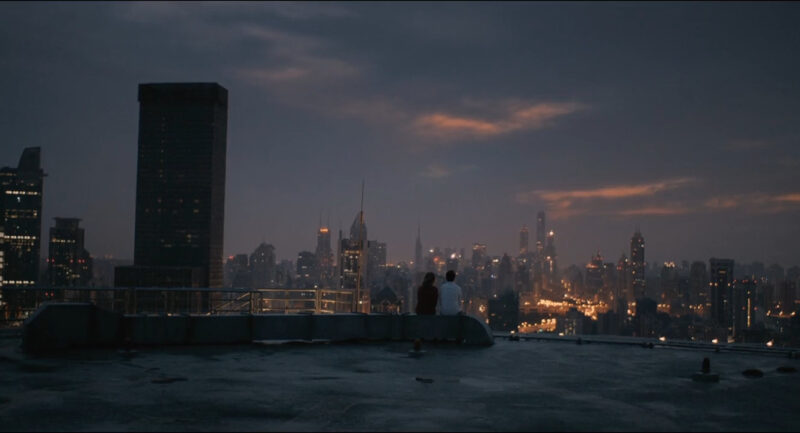 A woman and a man sit on a rooftop and watch a city at night
