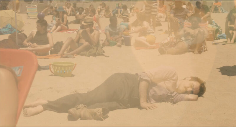 A man lies on a beach populated by many other people fully clothed