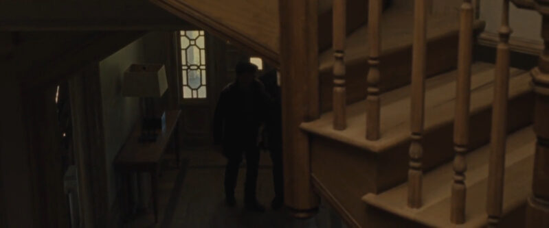 Two men stand in the dark downstairs with a staircase to their right