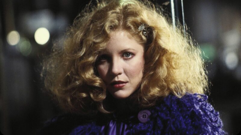 A blonde woman with curly hair wearing purple stares intently