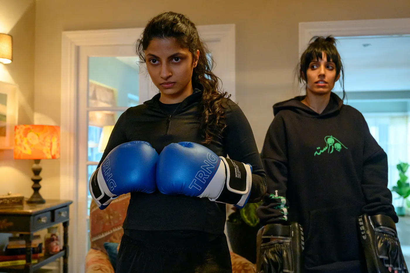 Ria and Lena Khan stand in a room. Ria has on boxing gloves.