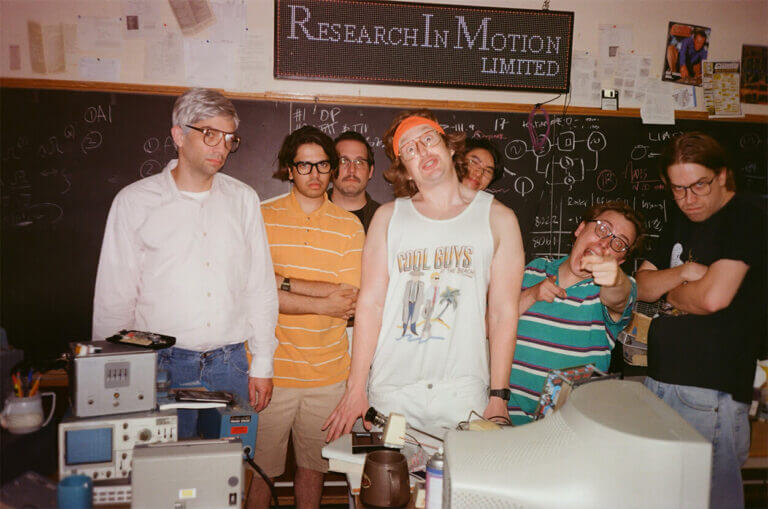 A group of nerdy guys stand in a computer lab in the mid 1990s.