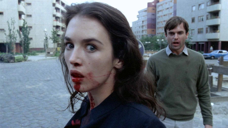 A woman with a bloody mouth looks at the camera while a man stands behind her
