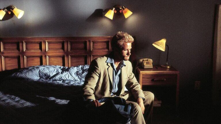 A blonde man in a tan suit sits on a bed with blue sheets
