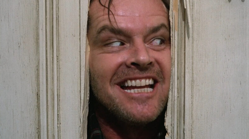 A man smiles manically as he sticks his head through a hole in a door