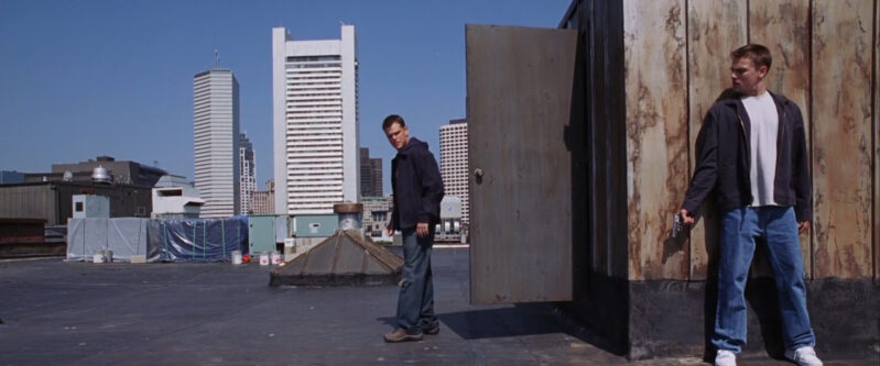 A man walks onto a roof as another man waits behind a corner holding a gun