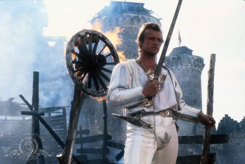A man looks down at his sword as a wheel burns behind him