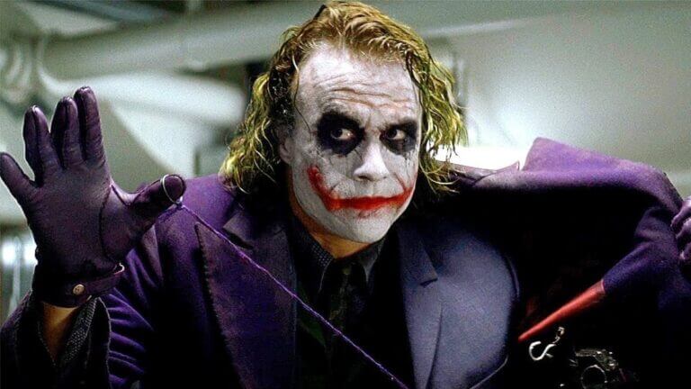 A man with clown makeup lifts his purple coat to reveal a grenade