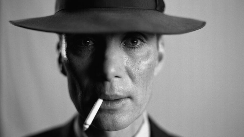 J. Robert Oppenheimer stares into the camera while wearing a hat and smoking a cigarette