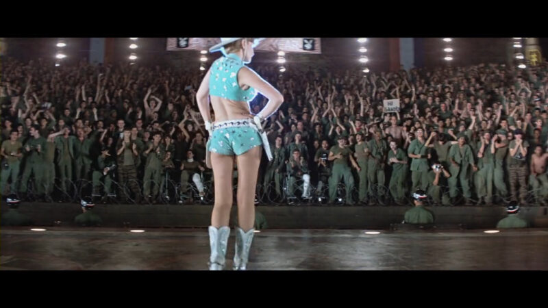 A woman in a dainty cowboy outfit stands before a crowd of soldiers