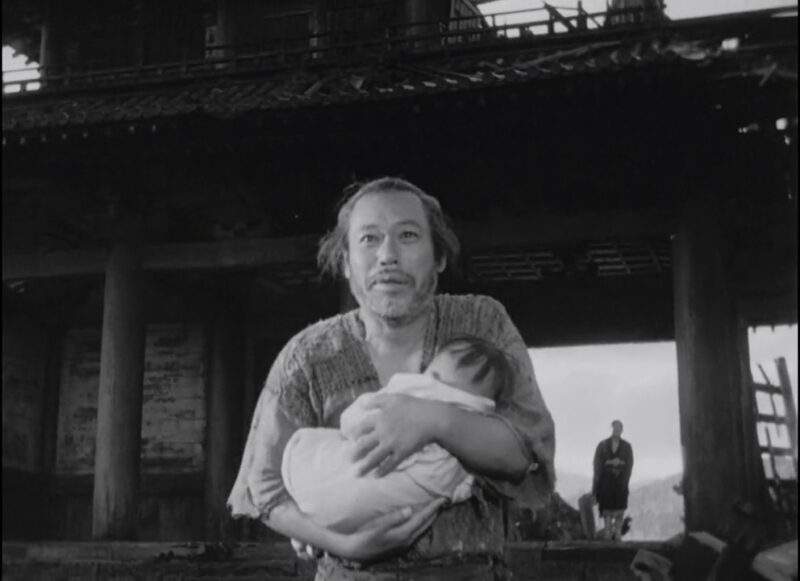 A man holds a baby as another man watches from the distance
