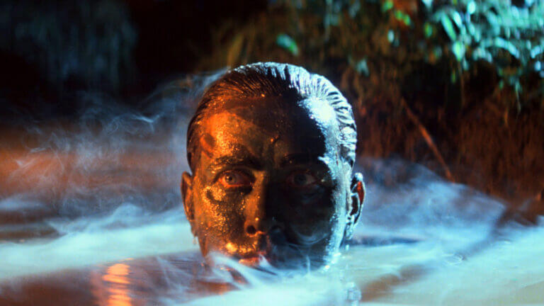 A man's face covered in army paint rises from the river in a jungle