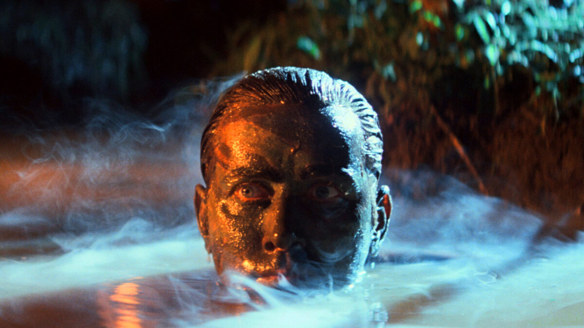 A man's face covered in army paint rises from the river in a jungle