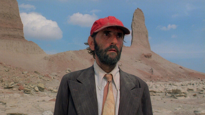 Travis stands in the desert wearing a suit and a red hat