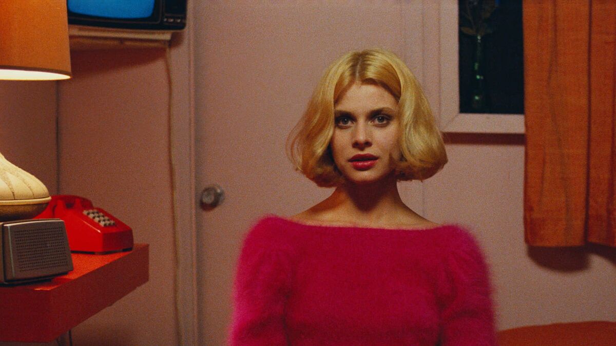 Jane, dressed in a pink sweater, sits in a pink peep show room