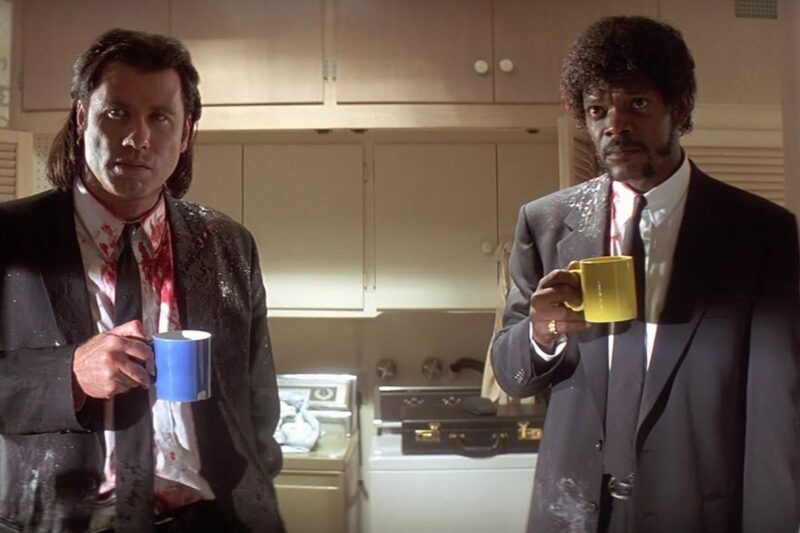 Vincent and Jules stand in a kitchen holding coffee mugs while wearing blood-stained suits