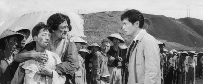 Two Chinese prisoners, surrounded by other prisoners, stare at Kaji in anger