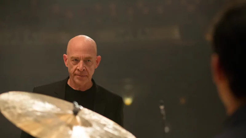 A man stares at another man who sits at a drum kit