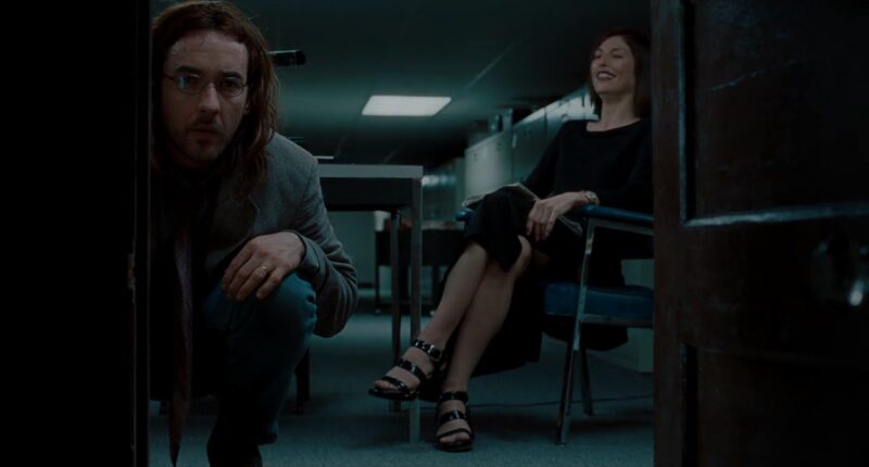 Maxine sits on a chair and smiles as Craig looks down a tunnel that leads to John Malkovich's mind