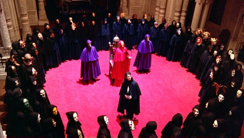 A group of cloaked and masked men surround Bill Harford on a red carpet as they look up towards the balcony