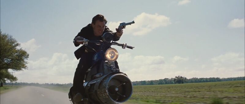 Kid Blue rides his motorcycle down a road in the cornfield pointing his gun