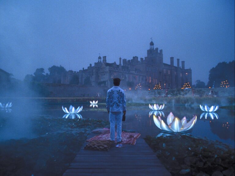 A man stands at the end of a dock, looking at an old English castle in the distance