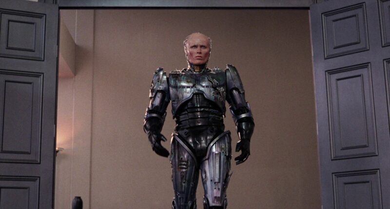 RoboCop, aka Murphy, walks into an OCP boardroom where a meeting is taking place